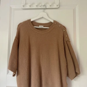Beige stickad tröja från Dante6 - Supermjuk beige stickad tröja från Dante6 med korta ärmar och snygga guldfärgade knappar på ena axeln. Tröjan har en loose fit och ribbade kanter vid hals, ärmslut och nederkant. Perfekt för lager-på-lager under hösten.