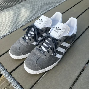 Adidas Gazelle grå/vita sneakers stl 44 - Snygga Adidas Gazelle sneakers i grå mocka med vita ränder och vit sula. Skorna har grå snörning, vit plös med Adidas-logga och diskret guldfärgad Gazelle-text på sidan. Använda 3 gånger och i väldigt bra skick. Bara att höra av sig vid eventuella frågor!😉