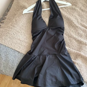 Svart kort halterneckklänning - Supersnygg svart kort klänning med shorts under kjolen, halterneck,  och v-ringning. Klänningen har en figurnära passform upptill och en lätt utsvängd kjol. Materialet är slätt och stretchigt, perfekt för en trendig look. Aldrig använt då den inte passar på mig