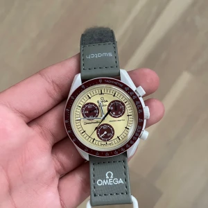Omega x Swatch MoonSwatch Pluto - Säljer en Omega x Swatch MoonSwatch Mission to Pluto. Klockan har en beige urtavla med vinröda detaljer och grått band med Omega- och Swatch-logga. Boetten är vit och klockan har kronograf-funktion. Perfekt för dig som gillar snygga och unika accessoarer.