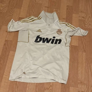 Real Madrid Ronaldo 9 vit fotbollströja - Säljer en vit Real Madrid fotbollströja från Adidas med guldiga detaljer och tre ränder på axlarna. Tryck med 'bwin' på framsidan och 'Ronaldo 9' på ryggen. Tröjan har korta ärmar, krage och är tillverkad i lätt, ventilerande material (Climacool). Perfekt för fans av Ronaldo!