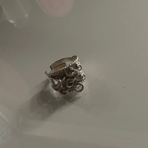 Silverring med hjärtdetalj (Kärleksringen) - Cool bred silverring med snirkligt mönster och en tydlig hjärtdetalj framtill. Ringen har en öppen baksida som gör den justerbar i storlek. Perfekt för dig som gillar statement-smycken med unika detaljer.