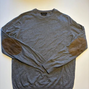 Grå Massimo Dutti tröja med mockadetaljer - Snygg grå Massimo Dutti i bomull, silke och kashmir. Tröjan har lång ärm och coola bruna mockalappar på armbågarna. Perfekt för dig som gillar klassisk stil med en twist. Rund halsringning och mjukt material. PRIS KAN DISKUTTERAS!