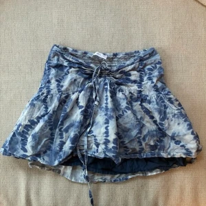 Blåvit kortkjol från MNG med snörning - Supersöt kortkjol från MNG i blått och vitt batikmönster. Kjolen har en volangig form och smock i midjan för extra bekväm passform. Snörning framtill ger en snygg detalj. Perfekt för varma dagar och enkel att styla med en enkel topp.