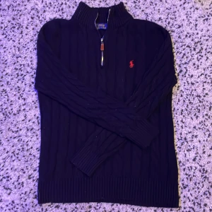 Svart stickad half zip från Ralph Lauren - Svart stickad tröja från Ralph Lauren med half zip och klassisk röd logga på bröstet. Tröjan har kabelstickat mönster, hög krage och långa ärmar. Perfekt för dig som gillar en stilren och tidlös look.