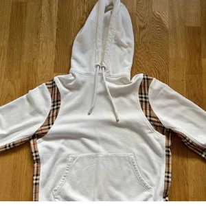Burbery  tröja  - Snygg vit hoodie från Burberry med klassiska beige, svarta och röda rutiga paneler längs sidorna och ärmarna. Tröjan har huva med snörning och en stor magficka framtill. Perfekt för dig som gillar streetstyle med lyxig touch. Den är äkta lappar finns. Pris diskuteras vid snabb affär 