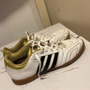 Adidas vita fotbollsskor med guld - Snygga vita Adidas fotbollsskor med klassiska svarta ränder på sidorna och gulddetaljer vid hälen. Skorna har bruna gummisulor, snörning och är tillverkade i skinn. Perfekta för inomhusspel och har en sportig vibe.