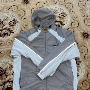 Grå och vit vindjacka från Corteiz - Säljer en snygg vindjacka från Corteiz i grått och vitt med reflekterande detaljer längs ärmarna. Jackan har huva med snörning, dragkedja framtill och coolt broderat märke på bröstet. Perfekt för dig som gillar streetwear och vill sticka ut.