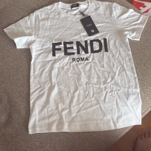 Vit Fendi t-shirt med tryck - Vit t-shirt från Fendi med svart FENDI ROMA-tryck på bröstet. Klassisk rund hals och korta ärmar. Mjuk bomullskvalitet som känns skön mot huden. Perfekt för dig som gillar stilrena designer och vill ha ett statement-plagg i garderoben.