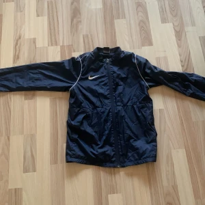 Mörkblå vindjacka från Nike - Snygg mörkblå vindjacka från Nike med hel dragkedja och vita detaljer längs axlarna. Jackan har lång ärm, hög krage och är tillverkad i lätt polyester som passar perfekt för aktiva dagar. Klassisk sportig look med Nike-logga på bröstet. Passar även bra på folk som är 170 cm 