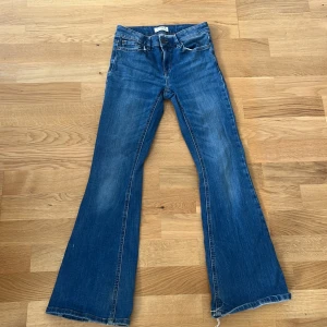 Blå bootcut jeans med rå kant - Säljer ett par klassiska blå jeans med bootcut passform och rå, fransad kant nertill. Jeansen har fem fickor, bälteshällor och dragkedjegylf. Perfekta för dig som gillar en retro vibe och vill ha ett par jeans med lite extra attityd.