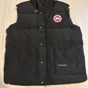 Svart dunväst från Canada Goose L - Svart dunväst från Canada Goose i storlek L. Västen har två stora fickor framtill, tryckknappar med logga och klassisk Canada Goose-patch på bröstet. Fylld med dun och har en hög krage för extra värme. Perfekt för kalla dagar och snygg till streetwear.