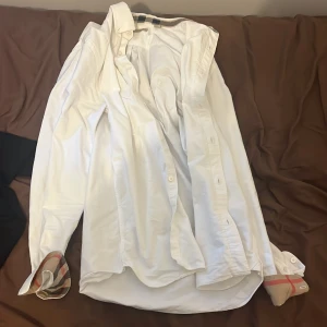 Vit Burberry Brit skjorta med rutiga detaljer - Vit skjorta från Burberry Brit i bomull med klassiska rutiga detaljer på insidan av kragen och manschetterna. Skjortan har lång ärm, knappar framtill och en stilren, tidlös look. Perfekt för dig som gillar exklusiva och snygga plagg med ikoniska detaljer.