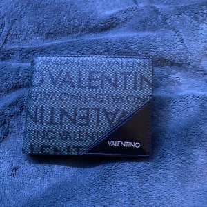 Valentino Svart Plånbok Helt ny - Snygg svart plånbok från Valentino med logomönster över hela utsidan och en stilren design. Plånboken har flera kortfack, myntficka och fönster för ID-kort. Tillverkad i skinn med detaljer i syntet och RFID-skydd. Perfekt för dig som vill ha något både praktiskt och trendigt. Den är helt ny och aldrig använd. Pris kan diskuteras!