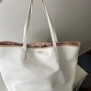 Vit shoppingväska från Don Donna - Stor och stilren shoppingväska från Don Donna i vitt skinnimitation. Väskan har långa handtag, ljusrosa kant och randigt innerfoder med dragkedjefack. Perfekt för att få plats med allt du behöver till skolan eller stan.