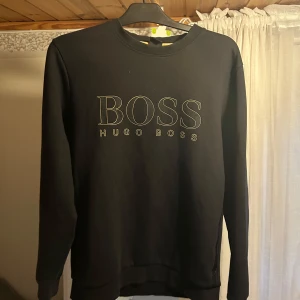 Svart sweatshirt från Hugo Boss  - Svart sweatshirt från Hugo Boss med stort BOSS-tryck framtill. Klassisk rund halsringning och långa ärmar. Tröjan är i mjuk bomull. Ett litet hål i nacken fingerstort
