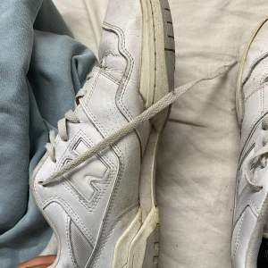 Vita New Balance 550 sneakers - Säljer ett par vita New Balance 550 sneakers med klassisk retrodesign. Skorna har snörning, perforerade detaljer på sidorna och en platt sula. Märkets logga syns tydligt på tungan och sidan. Perfekt för dig som gillar stilrena och sportiga sneakers.