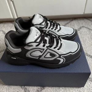 Dior B30 sneakers i svart och grå - Snygga Dior B30 sneakers i svart och grå mesh med stora CD-loggor på sidorna. Skorna har svarta snören, rund tå och en chunky platt sula. Kombinationen av mesh och syntet ger en modern vibe och detaljerna gör dem till ett statement-piece för din outfit.