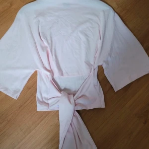 Ljusrosa omlottopp med knyt - Supersöt ljusrosa omlottopp med knyt i midjan och trekvartsärmar. Toppen har en avslappnad passform och är gjord i mjuk bomull, perfekt för en chill och trendig look.  Har MASSOR av kläder på min v1nted, vill du kika på fler plagg så skicka pm så skickar jag länk 🍒