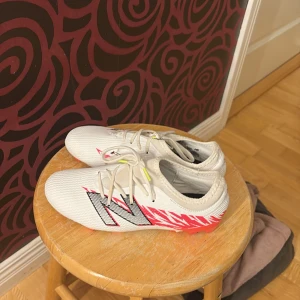 Vita New Balance Furon fotbollsskor - Säljer ett par vita New Balance Furon fotbollsskor med snygga rosa och svarta detaljer. Skorna har snörning och en lätt ovandel i syntetmaterial för bra känsla på planen. Sulan är rosa och vit med fasta dobbar, perfekt för gräsplaner. Stora NB-loggor på sidorna.