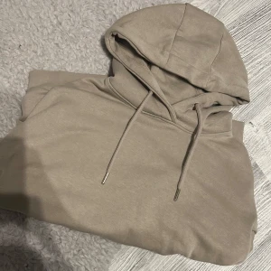 Beige hoodie från RYVLS med huva - Snygg beige hoodie från RYVLS med klassisk huva och dragsnören. Tröjan har en stor magficka och ribbade muddar vid ärmslut och nederkant. Perfekt för chill dagar och enkel att matcha med jeans eller joggers.