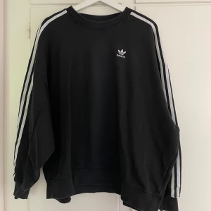 Svart Adidas sweatshirt med vita ränder - Svart oversized sweatshirt från Adidas med klassiska vita ränder längs ärmarna och broderad logga på bröstet. Vet ej fraktkostnaden.