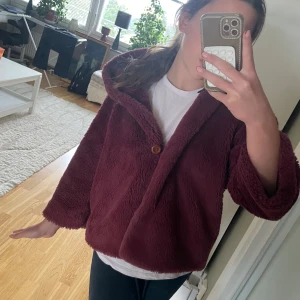 Vinröd teddyjacka med huva - Mysig vinröd teddyjacka med trekvartsärm. Jackan har en loose passform och är tillverkad i fluffigt teddy-material som håller dig varm. Perfekt för dig som vill ha en chill och trendig look under kyliga dagar.