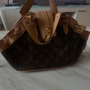  handväska - Säljer en klassisk handväska i brunt skinn med ikoniskt monogrammönster och ljusbruna detaljer. Väskan har guldfärgade metalldetaljer, lås och dragkedja samt rymligt foder inuti. Perfekt för dig som vill ha en lyxig och tidlös accessoar.