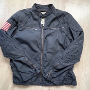Svart army jacka Ralph Lauren Denim & Supply - Svart jacka från Ralph Lauren Denim & Supply med klassiska ralph lauren amerikanska flag patch på ärmen. Jackan har ståkrage, dragkedja framtill, två bröstfickor med dragkedja och quiltade detaljer på axlar och ärmar.  Storlek XL men sitter som L                                                                         Pris diskuterbart