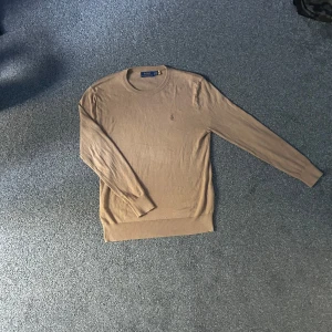 Beige tröja från Polo Ralph Lauren - Snygg beige tröja från Polo Ralph Lauren med rund halsringning och diskret broderad logga på bröstet. Långärmad modell i mjukt material som sitter skönt och har ribbade muddar vid ärmslut och nederkant. Perfekt för en clean och stilren look.