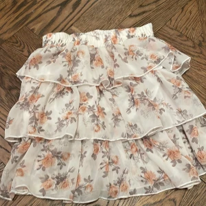 Blommig volangkjol från Happy Star -  kort volangkjol från Happy Star med blommigt mönster i beige, grått och persikofärgat på vit botten. Kjolen har flera lager volanger och resår i midjan. Storlek 140 cm