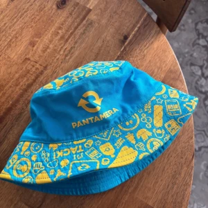 Pantamera bucket hat - Helt ny pantamera bucket hat.  Här är en rolig och färgglad Pantamera bucket hat. Den har ett unikt mönster med Pantamera-logotyper och andra återvinningsrelaterade symboler i gult tryck på en turkos bakgrund. Perfekt för att visa ditt stöd för miljön med stil!  One size.