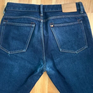 Mörkblå straight jeans från & Denim - Snygga mörkblå jeans från A Denim med klassisk femficksdesign och raka ben. Jeansen har kopparfärgade nitar och kontrastsömmar, samt en läderpatch bak i midjan. Perfekta för dig som gillar en tidlös och clean look. Ej stretch.