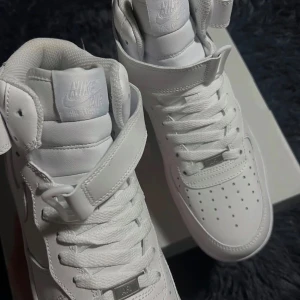 Nike Air Force 1 Mid Triple White 42 - Nike Air Force 1 Mid i helvitt skinn med klassisk snörning och kardborreband vid ankeln. Snygg och clean siluett med perforerad tåbox och robust platt sula. Perfekt för dig som gillar tidlös streetstyle och vill ha ett par fräscha sneakers i garderoben.