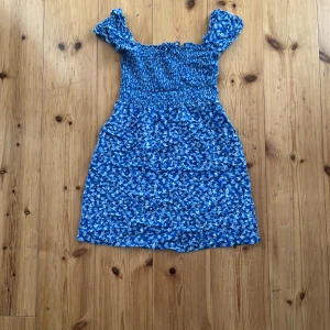 Blå blommig klänning New Yorker - Supersöt blå kortklänning från FB Sister i storlek XXS. Klänningen har blommigt mönster, smockad överdel och korta puffärmar som kan bäras offshoulder. Perfekt för varma dagar och ger en fräsch sommarkänsla.