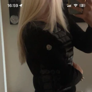 Svart Moncler cardigan jacka - Snygg svart cardigan jacka från Moncler med quiltad framsida och ärmar i stickad ull. Jackan har dragkedja framtill, två fickor med knapp och Moncler-logga på ärmen. Perfekt för dig som vill ha en stilren och sportig look.