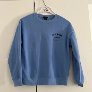 Blå sweatshirt från new yorker  - Ljusblå sweatshirt från new yorker i storlek XS. Tröjan har rund hals, tryck med texten 'ESSENTIAL COLLECTION' på bröstet och långa ärmar. Mjuk insida och ribbade muddar vid ärmslut och nederkant