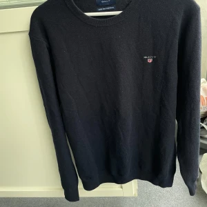 Mörkblå stickad tröja från Gant - Mörkblå stickad tröja från Gant med rund halsringning och broderad logga på bröstet. Tröjan är i superfin lammull och har långa ärmar. Perfekt för dig som gillar klassisk och stilren look.