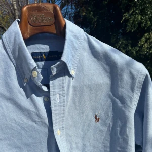 Ljusblå skjorta från Ralph Lauren - Klassisk ljusblå skjorta från Ralph Lauren med button-down krage och broderad logga på bröstet. Skjortan har långa ärmar och knäppning framtill. Perfekt för en clean och stilren look.