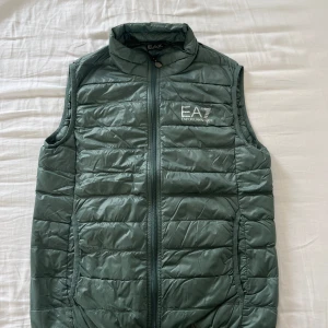 Grön dunväst från Emporio Armani EA7 - Snygg grön dunväst från Emporio Armani EA7 med quiltad design och dragkedja framtill. Västen har hög krage och två fickor på sidorna. Perfekt att slänga över en hoodie för en chill och sportig look.