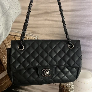 Svart quiltad axelväska  - Säljer en ikonisk svart axelväska med klassiskt quiltat mönster och silverfärgat CC-spänne. Väskan har en kedjerem med insytt skinn och lockstängning. Perfekt för dig som vill ha en tidlös och lyxig accessoar.