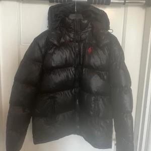 Svart dunjacka från Polo Ralph Lauren - Snygg svart dunjacka från Polo Ralph Lauren med röd logga på bröstet. Jackan har en glansig finish, huva och dragkedja framtill. Perfekt för kalla dagar och riktigt skön att bära. Klassisk pufferstil som alltid är rätt.