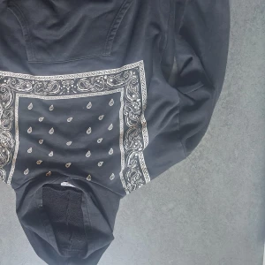 Svart hoodie med paisleymönster - Svart hoodie från Fashion Woman med vit paisleyinspirerad bandana-print på framsidan. Tröjan har huva och är fodrad med mjukt material på insidan. Perfekt för dig som gillar streetstyle och vill sticka ut lite extra.