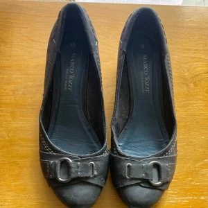 Svarta ballerinaskor från Marco Tozzi - Snygga svarta ballerinaskor från Marco Tozzi med rund tå och låg kilklack. Skorna har dekorativa spännen framtill och är gjorda i skinn med synliga sömmar längs kanterna. Perfekta för dig som gillar klassisk och stilren look. Av använda skick, köpta på loppis 2025 för 100kr.