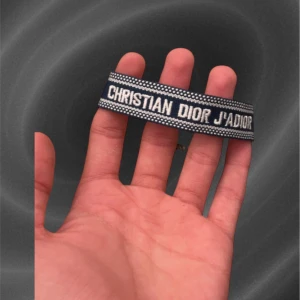 Blått Christian Dior J'adior armband - Snyggt blått armband från Christian Dior med texten 'J'adior' broderad i vitt. Bandet har en randig design och är tillverkat i ett mjukt tygmaterial. Perfekt accessoar för att lyfta din outfit med lite lyxig vibe.