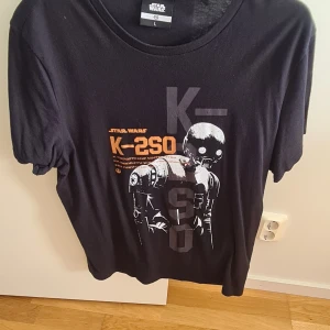 Star Wars K-2SO svart t-shirt - Svart t-shirt med tryck av K-2SO från Star Wars på framsidan. Snygg orange och grå text och robotmotiv. Klassisk passform med rund hals och korta ärmar. Tillverkad i mjuk bomull, perfekt för dig som älskar sci-fi och coola prints.