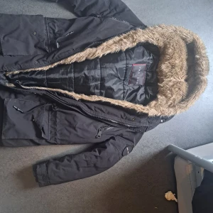 Svart parkajacka med fuskpäls från Pegador - Svart parkajacka från Pegador i storlek medium. Jackan har en stor huva med brun fuskpäls, flera fickor med dragkedja och snörning samt quiltad insida. Perfekt för kalla dagar när du vill hålla stilen och värmen.