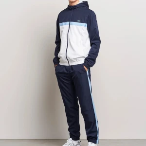 Blå och vit träningsdress från Lacoste - Snygg träningsdress från Lacoste i mörkblått och vitt med ljusblå detaljer. Jackan har huva, dragkedja och elastiska muddar. Byxorna har en ljusblå rand längs sidorna och resår vid benslut. Perfekt för dig som gillar sportig stil.