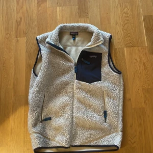 Beige fleeceväst från Patagonia - Säljer en beige fleeceväst från Patagonia med marinblå detaljer och bröstficka. Perfekt nu till hösten!