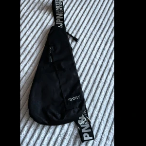 Svart sportig axelväska med tryck - Säljer en svart axelväska i syntetmaterial med justerbart band med vit text och coolt tryck. Väskan har dragkedja och en sportig look, perfekt för att bära det viktigaste. Enkel och stilren design med tydlig 'SPORT'-detalj framtill.